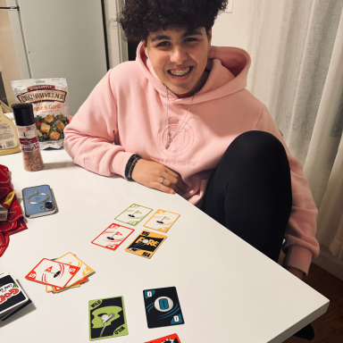A person wearing a pink hoodie sits at a table with playing cards and snacks.